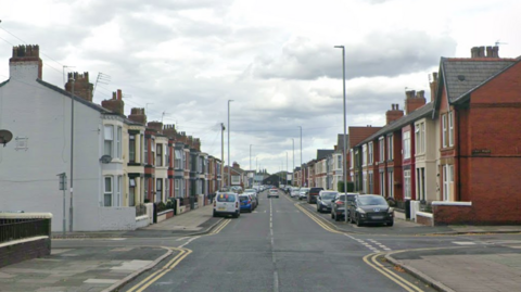 A road is lined with terraced houses and cars are parked at either side of the road. A road leads off to the left and there is one leading off to the right.