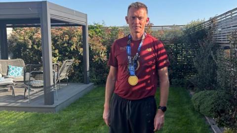 Danny Stapleton in his garden with a medal round his neck, dressed in a red t-shirt and black shorts