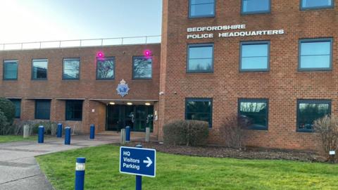 The front of Bedfordshire Police headquarters. It is a red brick building with grass and bushes outside. In the foreground there is a sign for visitors parking.
