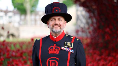 Andy Harris in full Yeoman Warder uniform. He wears a tall navy hat, a navy jacket with a red collar. 