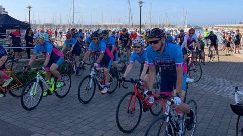 A picture of cyclists all on their bikes ready for a race. A majority of them are wearing a blue and pink outfit. The pier can be seen in the background.