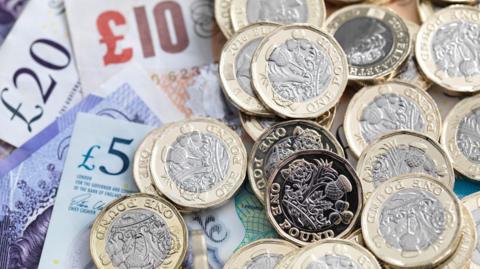 UK Sterling £5, £10 and £20 notes lie on top of one another with a handful of golf and silver pound coins lying on top of the notes.