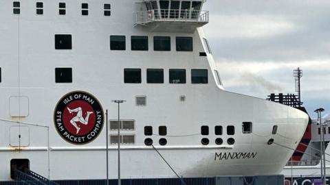A close up of the bow of Manxman, it is a large boxy white ferry, with a logo reading Isle of Man Steam Packet and the three legs of man.