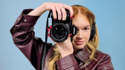 Girl wearing a maroon leather jacket is holding a black Nikon Z8 camera with a large lens and visible lens markings. The camera has a black strap hanging down and is positioned close to the person’s head. Her hand, with long red nails and a silver ring, is gripping the top of the camera