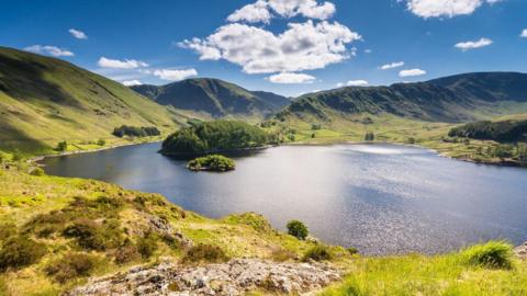 Aerial view of Haweswater. The reservoir is ringed by mountains, green and grassy in the sunlight, which is also reflected on the surface of the water. A promontory extending into the water is covered with conifers, and there are further patches of conifers to either side.
