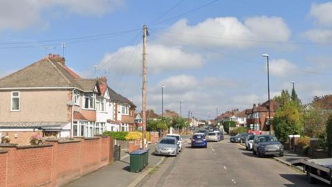 William Bristow Road has houses on both sides of the street and parked cars. There are bins, trees and lamp posts and the junction with Daventry Road is ahead.