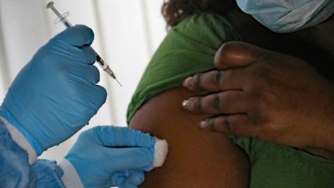A person wearing gloves - holding a needle and a cotton - giving an injection to a lady -in a green T-shirt.