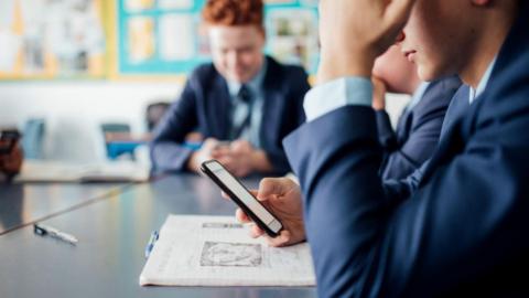 Boy using a smartphone in a classroom. Anonymised photo so you can only see his arms leaning on a desk and he's looking downwards at the phone screen. Behind him is another boy looking at his phone.