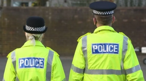Two police officers stand with their backs to the camera in uniform and florescent jackets. One officer is female and the other officer is male
