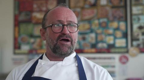 Daniel Smith inside a classroom wearing chef whites and a blue apron. He has a pair of black framed circular glasses on and is looking off camera mid-speech.
