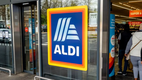 The front entrance to a branch can be seen with the shop's logo on a glass door and shoppers inside.