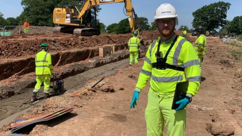Chris Rodgers pictured at a construction site. He is wearing high vis construction gear and a hard hat. Other workers and a digger can be seen in the background.
