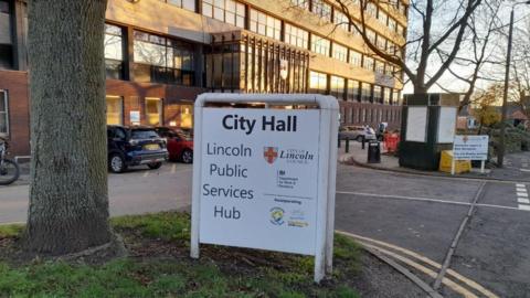 A sign outside a large office building labelled "City Hall." The sign reads "Lincoln Public Services Hub" and lists various public services. There are several parked cars and trees nearby.
