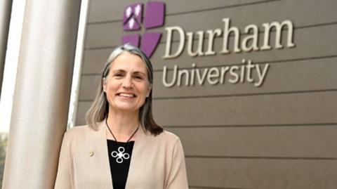 Fiona Hill, Durham University Chancellor, outside the a university building standing in front of a Durham University sign. She has shoulder-length grey hair and is wearing a pink blazer over a black top, with a large necklace with a metal pendant.