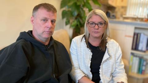 Jenny and Allyn Condon sit together on a sofa looking at the camera. They both have very serious expressions on their faces. He is wearing a black hooded top and she has a white cardigan and black top on