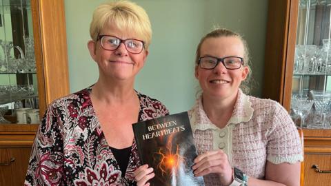 A mother and daughter, both wearing glaseses, sat down with a book called Between Heartbeats. The mother has blonde hair and a purple, grey and black patterned top. Her daughter is wearing a pink and white short-sleeved top.