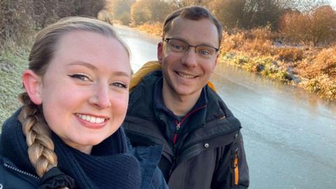 Alexandra Petri and Ross Jones wearing winter coats and standing beside a frozen river