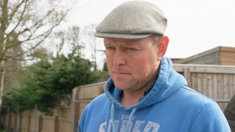 A clean shaven man is wearing a grey flat cap and a sky blue hoodie. He is standing in front of a wooden fence.