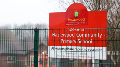 A red and white sign sits behind a green metal fence. The sign reads 'Welcome to Hazlewood Community Primary School. All visitors report to reception. A brick building and trees can be seen behind the sign.