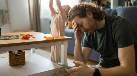 A man sitting on the floor of a living room and holding a phone in one hand with his other hand on his forehead. He is looking down towards his phone. In the background is a toddler playing. 