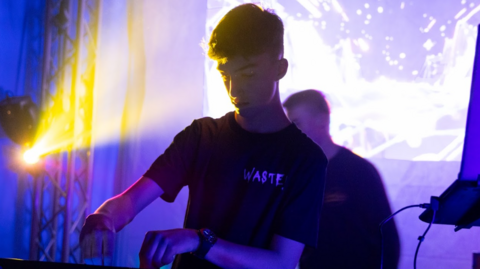Sam is mixing tracks at a club night. He is wearing a black t-shirt with the word 'Wasted' on the left hand side. He is standing in front of a screen projecting rave patterns.