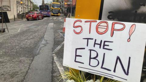 A sign which says "STOP! THE EBLN" positioned a graffitied road sign on a main road in Barton Hill. Two buses and a red car can be seen driving down the road.