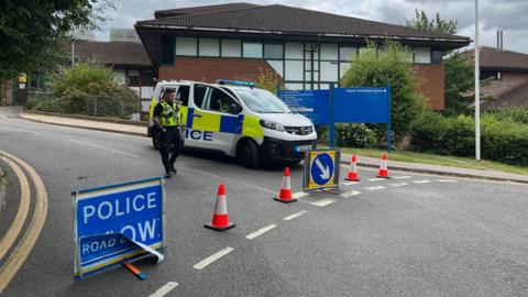 A police car parked and officer stood behind a row of orange-neon traffic cones and a sign reading "road closed".