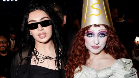 Charli XCX and Chappell Roan at the Grammy's. Charli wears all black including her signature black sunglasses, her curly black hair worn loose. Chappell wears a pointed gold had and a pale sea-green dress. She has curly red hair worn loose and wears extravagant makeup including pencil thin brows, white foundation and pink and blue eyeshadow. 