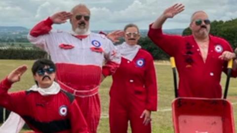 Four Red Barrows, in red costumes and sunglasses and moustaches. They are standing on green grass. There are green trees behind them. They are posing. 