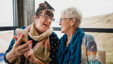 Two women laughing with each other. One is holding up a phone