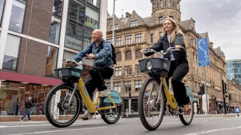 A man in a denim jacket cycles on a yellow bike alongside a woman in a black jacket who is also cycling nearby. The two are passing through a busy high street.