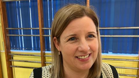 Clare O'Loan smiling at the camera, she has shoulder length blonde hair and blue eyes. She's wearing a black and cream patterned top, dropped earrings and standing in a school hall. 