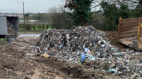 A lorry is parked on the left and the rear is open showing lots of rubbish inside. Piles of waste are also left on the side of the road or field.