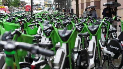 A pavement and road is filled with hundreds of parked, bright green Lime e-bikes. A few people with umbrellas are walking on the side.