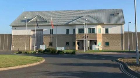 The exterior of the front entrance of Jurby Prison on a sunny day. The building is brown and cream in colour, has a grey pitched roof and flag poles flying the red Manx flag and signage in front of it. A tarmacked road leads up to it with grass on either side, and an a high perimeter fence can be seen around the complex. 