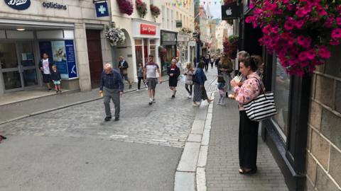 Guernsey's St Peter Port Town High Street. A woman is standing drinking a milkshake under a hanging basket of purple flowers. On the cobbled street, people are walking to and from the camera vantage point. Between the buildings on either side of the cobbled street is multi-coloured bunting. 