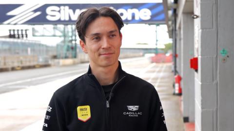 Driver Jack Aitken stands in a Cadillac Hertz Team Jota branded jacket in front of the Silverstone Circuit logo in an empty pit lane.