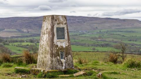 Thorny Gale Trig Pillar, which features a bronze plaque, standing on the top of a hill. A green valley and fells are in the background.