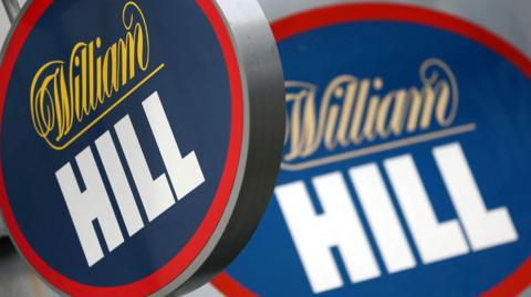 Two signs with the bookmaker William Hill logo on them. The one in the foreground is clearer and the one in the background is slightly blurry.