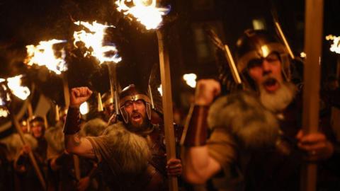 Men dressed in Viking gear carry flaming torches through the darkness