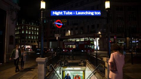 Entrance to a London Underground station at night, illuminated by streetlights and a blue sign reading “Night Tube - Launching tonight.” People walk near the station entrance, with city buildings, traffic, and a red double-decker bus visible in the background.