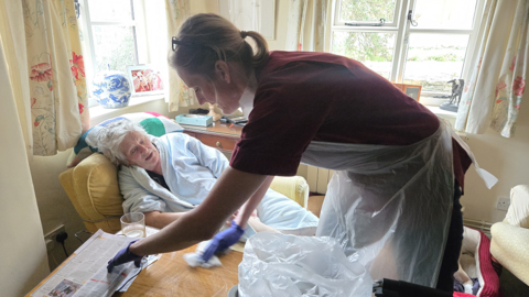 An elderly person in a dressing gown sits in an armchair in a living room. A nurse in red scrubs and a plastic apron is cleaning the area.