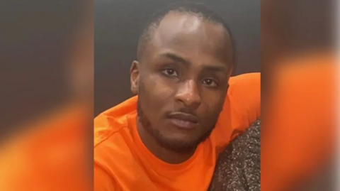 A man looking at the camera with a straight face, wearing an orange T-shirt. He has dark skin, a short beard and a mostly shaven head with very short, dark hair.