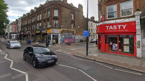 Street view image of the junction of Brixton Road and Handforth Road in Brixton. It is daylight and there are shops, takeaways and cars in sight