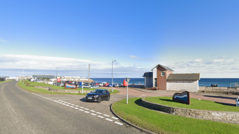 Road, with a black car at junction, and sea and blue sky in background