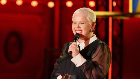 Comedian Laura Smyth, with cropped blonde hair holding a mic and smiling.