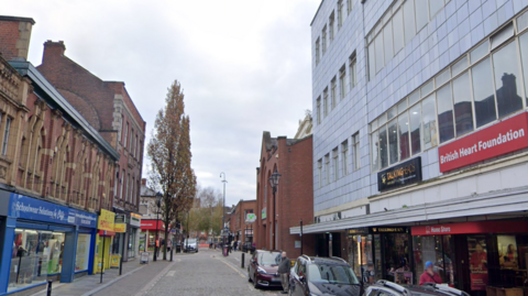 Printing Office Street in Doncaster city centre, with shops on both sides and cars parked down one side of the road.