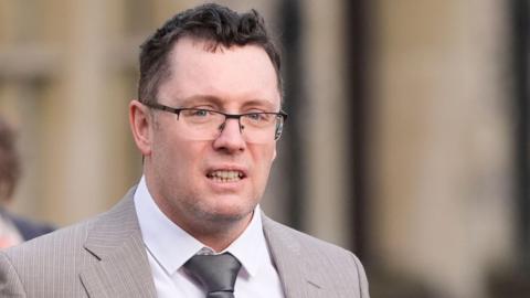 Christopher Trybus is walking outside a court in a grey suit with a white shirt. He has glasses and short black hair.