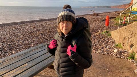 Sea swimmer Sarah Giles is standing on Budleigh Salterton beach. She is wearing a black coat, pink gloves and a brown, white and blue stripey woolly hat with a bobble. She is standing next to a wooden bench. The beach is pebbly. There are cliffs in the distance.