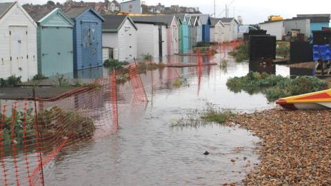 Beach huts surrounded by an overflow of water. Some pebbles can be seen on the right of the image 